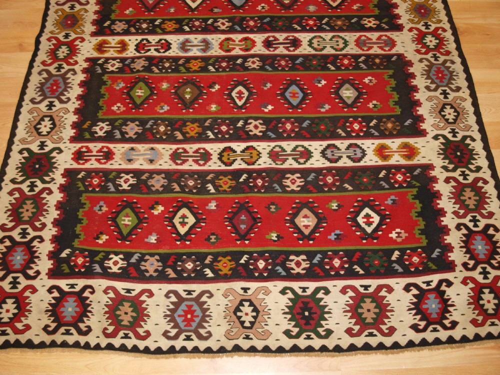 A rectangular handwoven antique wool kilim rug featuring horizontal red and dark brown banded panels decorated with geometric diamond motifs, framed by a broad cream border. View VI