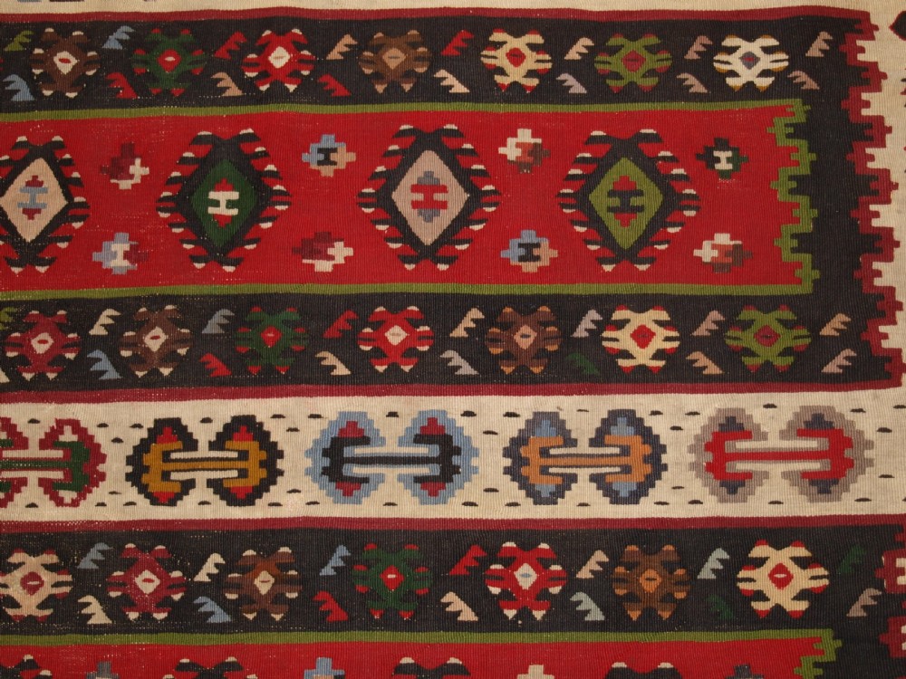 A rectangular handwoven antique wool kilim rug featuring horizontal red and dark brown banded panels decorated with geometric diamond motifs, framed by a broad cream border. View VII