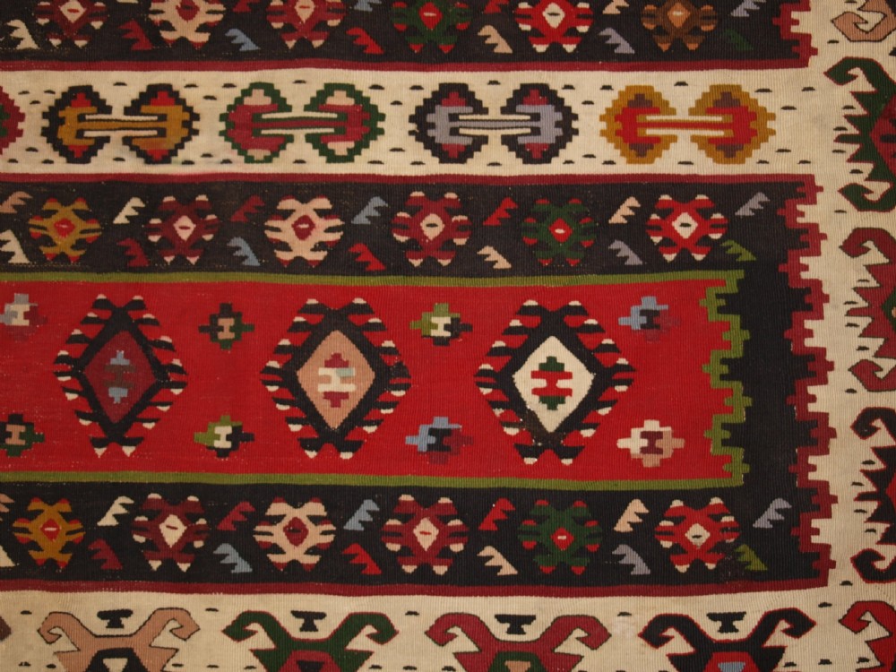 A rectangular handwoven antique wool kilim rug featuring horizontal red and dark brown banded panels decorated with geometric diamond motifs, framed by a broad cream border. View IX