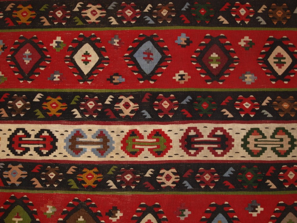 A rectangular handwoven antique wool kilim rug featuring horizontal red and dark brown banded panels decorated with geometric diamond motifs, framed by a broad cream border. View X