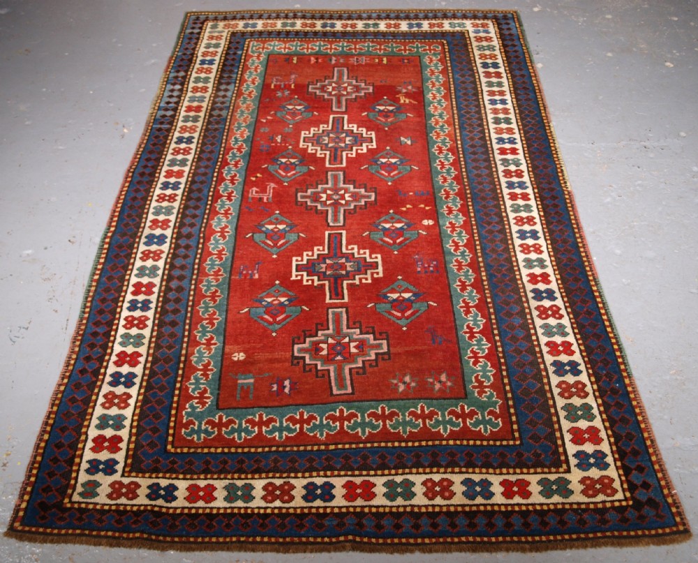 An antique Caucasian rug featuring a red field woven with five stacked geometric medallions, small animal figures, and multi-coloured patterned borders.