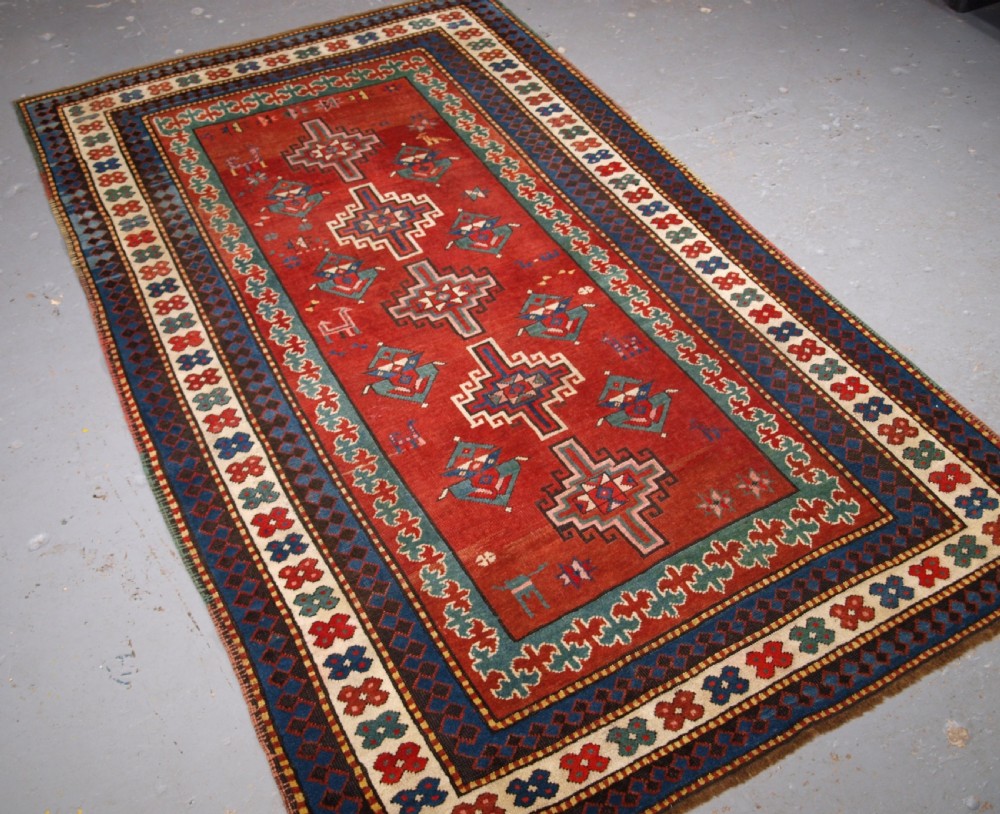 An antique Caucasian rug featuring a red field woven with five stacked geometric medallions, small animal figures, and multi-coloured patterned borders. View II