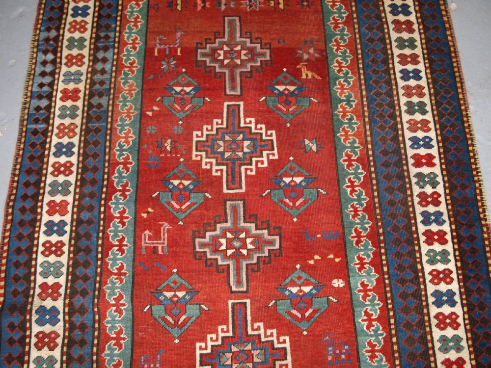An antique Caucasian rug featuring a red field woven with five stacked geometric medallions, small animal figures, and multi-coloured patterned borders. View III