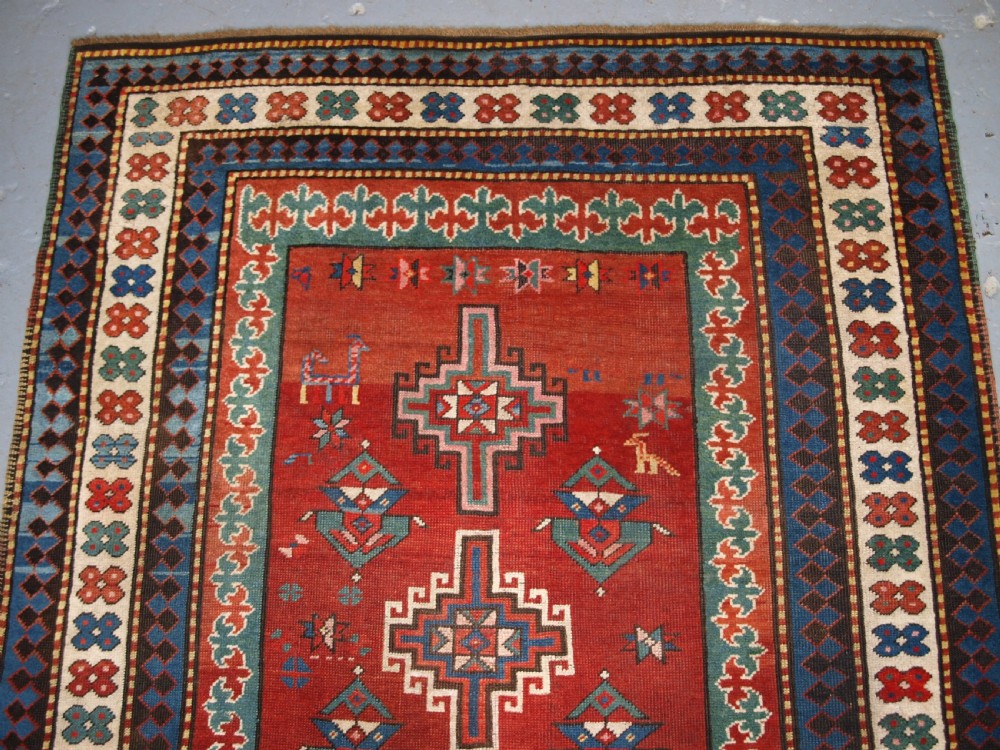 An antique Caucasian rug featuring a red field woven with five stacked geometric medallions, small animal figures, and multi-coloured patterned borders. View IV