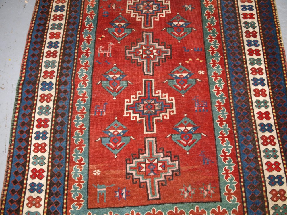 An antique Caucasian rug featuring a red field woven with five stacked geometric medallions, small animal figures, and multi-coloured patterned borders. View V