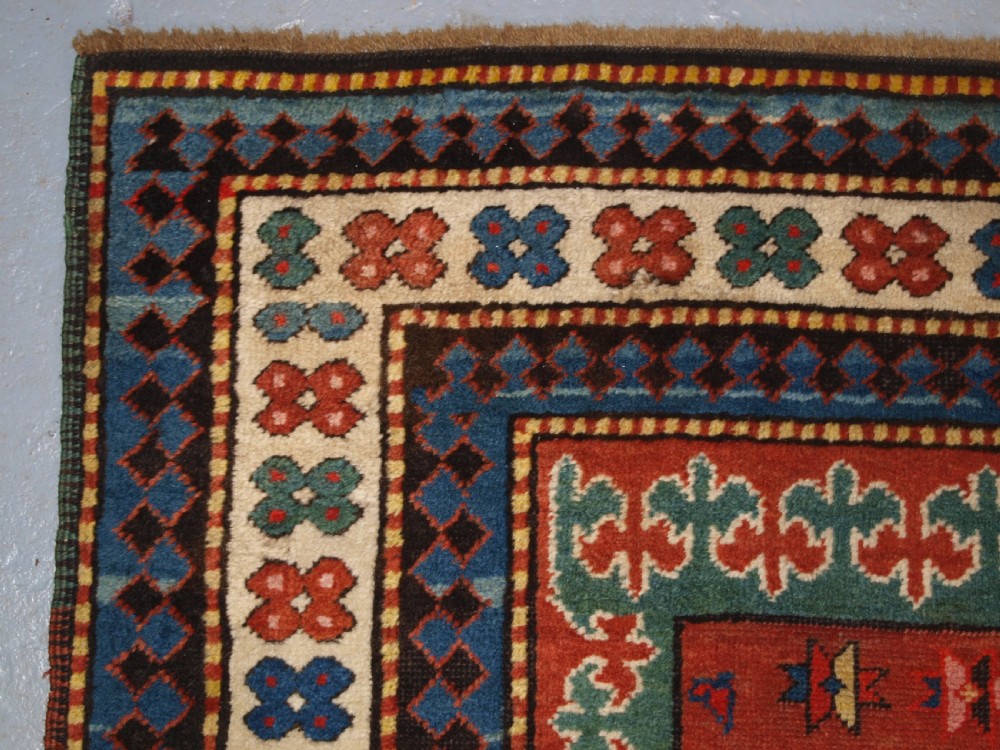 An antique Caucasian rug featuring a red field woven with five stacked geometric medallions, small animal figures, and multi-coloured patterned borders. View VII