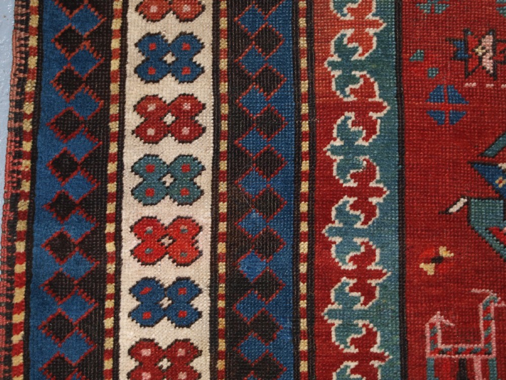 An antique Caucasian rug featuring a red field woven with five stacked geometric medallions, small animal figures, and multi-coloured patterned borders. View VIII