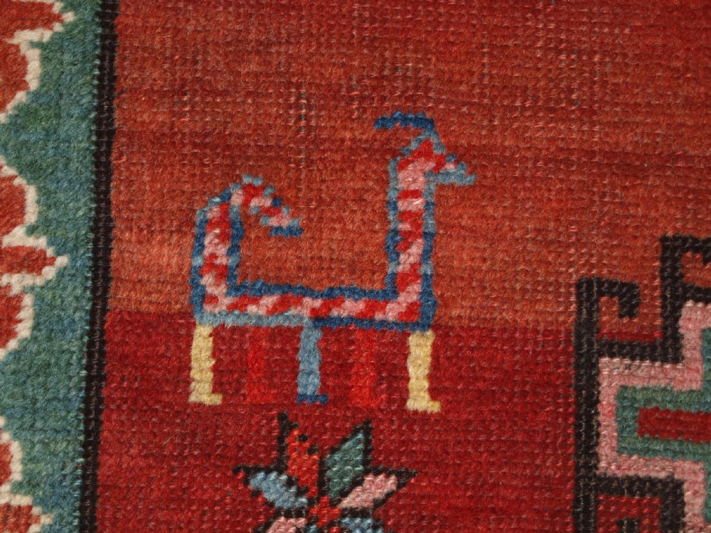 An antique Caucasian rug featuring a red field woven with five stacked geometric medallions, small animal figures, and multi-coloured patterned borders. View IX