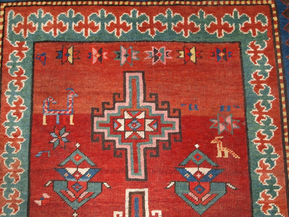 An antique Caucasian rug featuring a red field woven with five stacked geometric medallions, small animal figures, and multi-coloured patterned borders. View X
