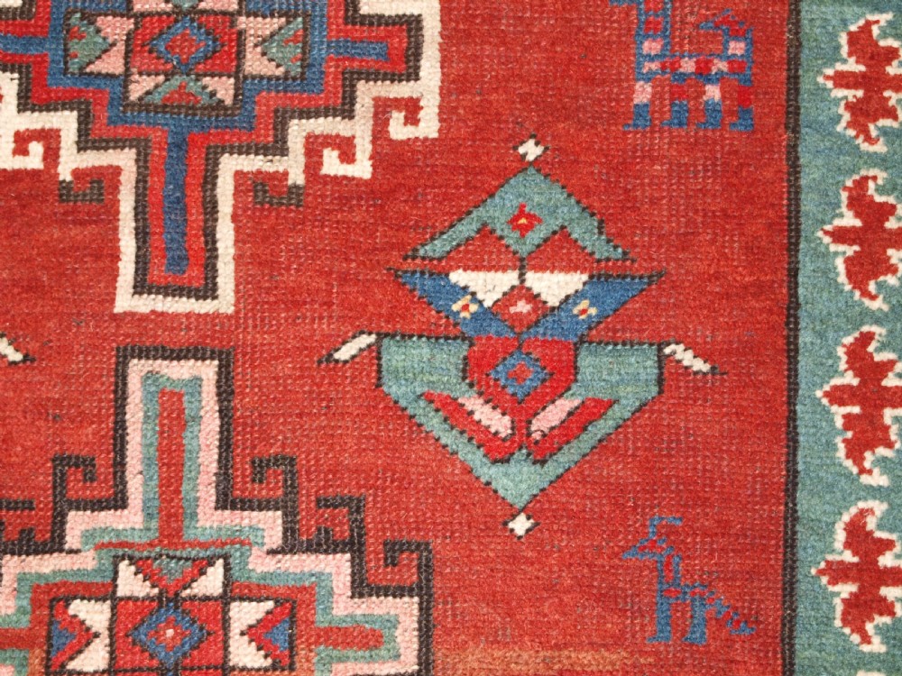 An antique Caucasian rug featuring a red field woven with five stacked geometric medallions, small animal figures, and multi-coloured patterned borders. View XI