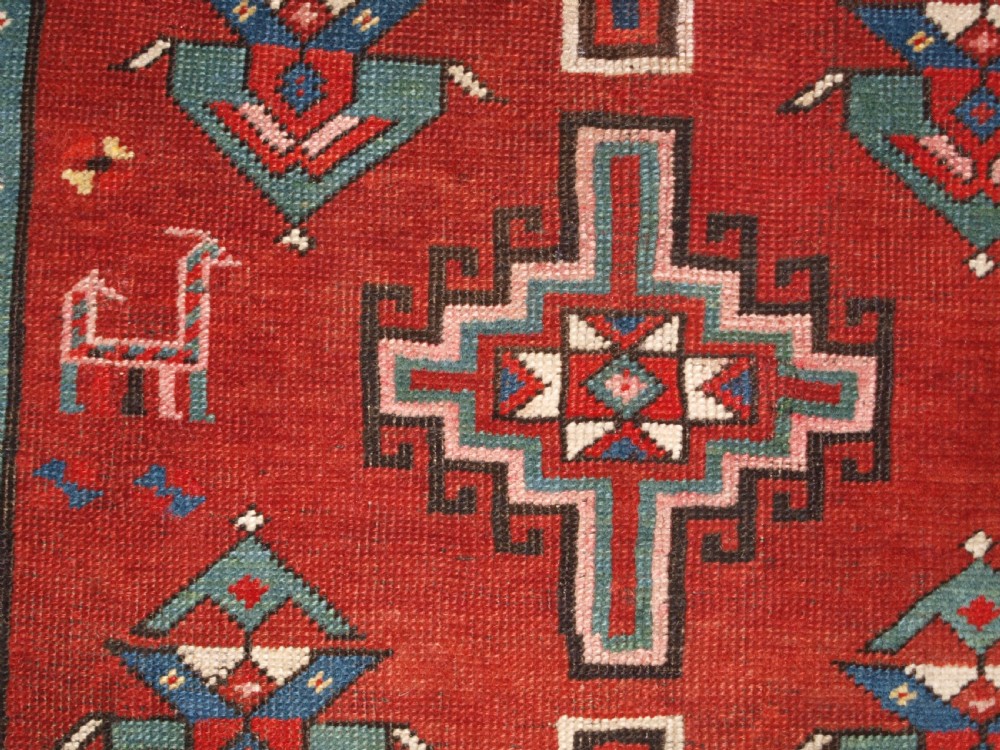 An antique Caucasian rug featuring a red field woven with five stacked geometric medallions, small animal figures, and multi-coloured patterned borders. View XII