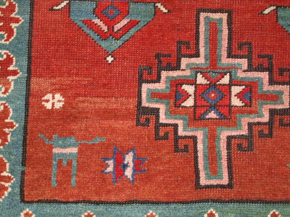 An antique Caucasian rug featuring a red field woven with five stacked geometric medallions, small animal figures, and multi-coloured patterned borders. View XIII