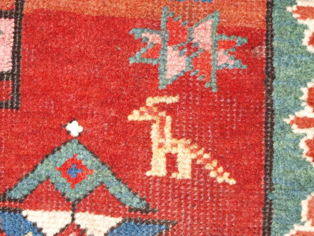An antique Caucasian rug featuring a red field woven with five stacked geometric medallions, small animal figures, and multi-coloured patterned borders. View XIV