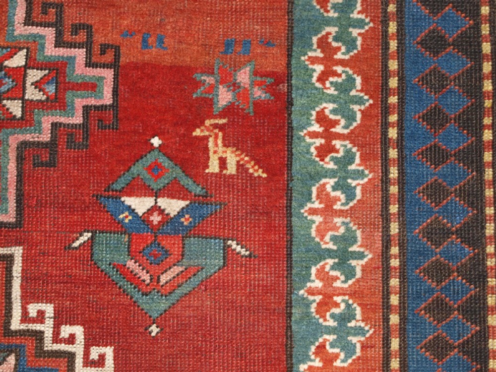 An antique Caucasian rug featuring a red field woven with five stacked geometric medallions, small animal figures, and multi-coloured patterned borders. View XV