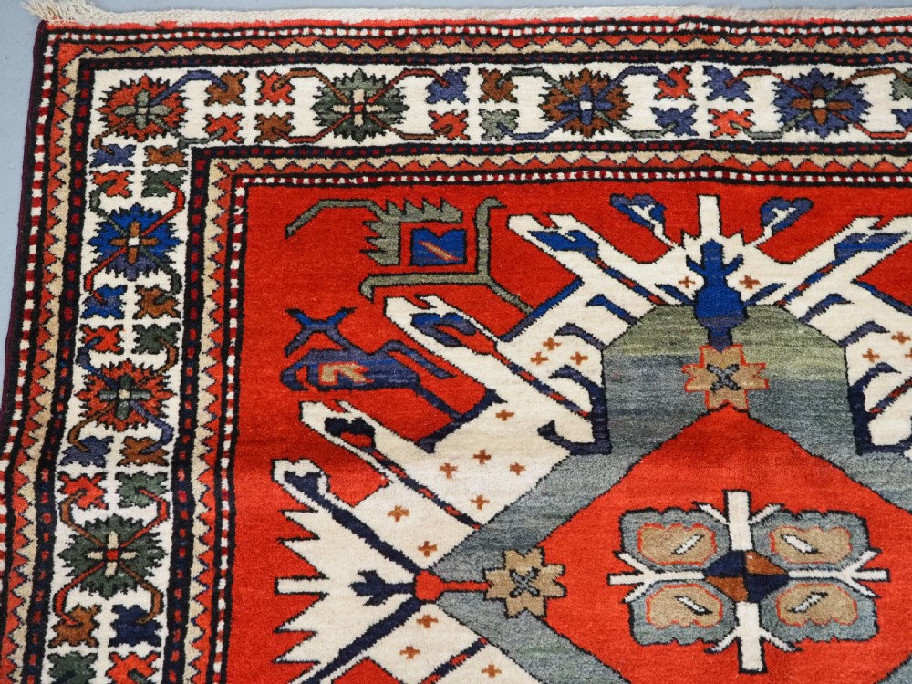 A hand-knotted Caucasian Eagle Kazak wool rug featuring radiating geometric medallions in cream, green, and navy set against a rich red central field. View VI