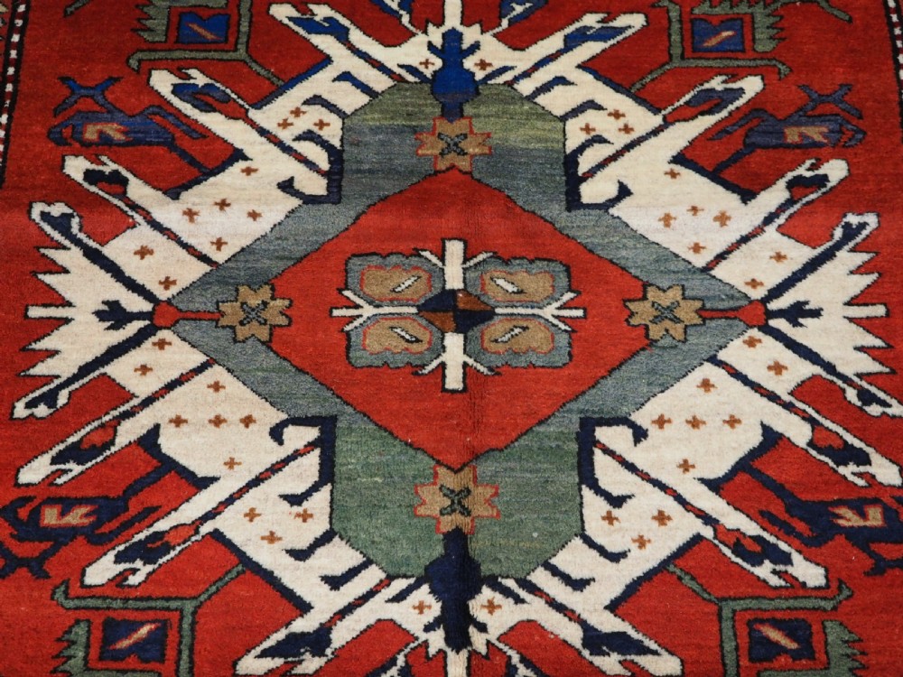 A hand-knotted Caucasian Eagle Kazak wool rug featuring radiating geometric medallions in cream, green, and navy set against a rich red central field. View VIII