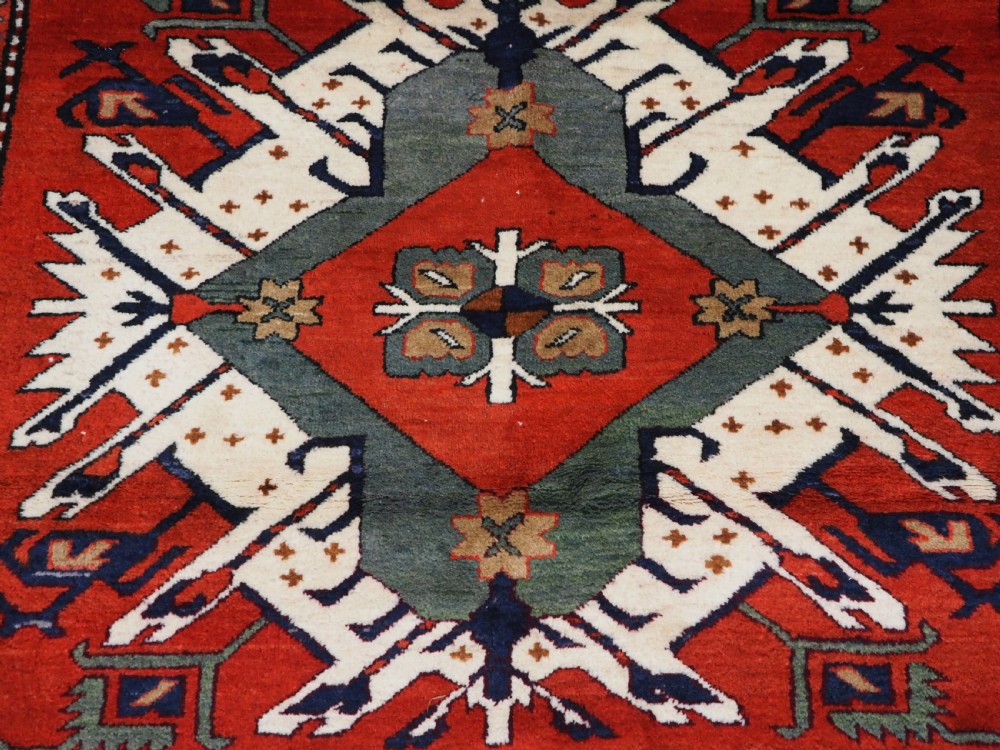 A hand-knotted Caucasian Eagle Kazak wool rug featuring radiating geometric medallions in cream, green, and navy set against a rich red central field. View IX