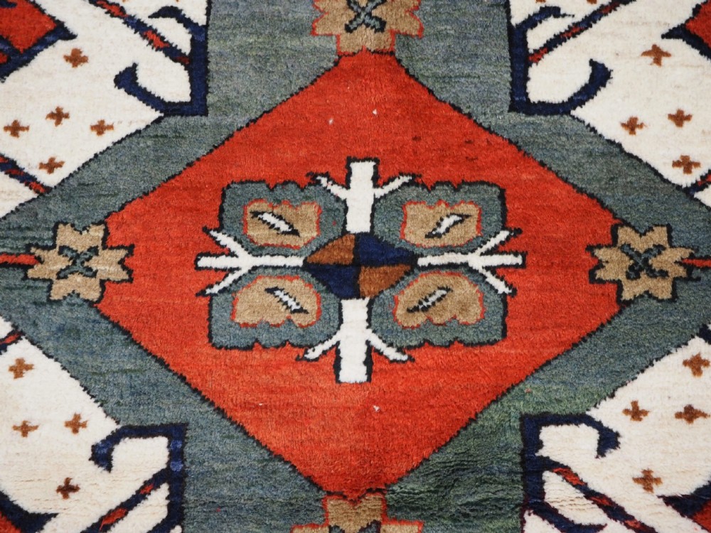 A hand-knotted Caucasian Eagle Kazak wool rug featuring radiating geometric medallions in cream, green, and navy set against a rich red central field. View XI