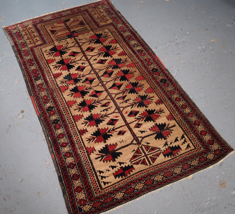 An antique Baluch wool rug featuring a central Tree of Life motif on a camel-coloured ground, framed by geometric red and black patterned borders. View II