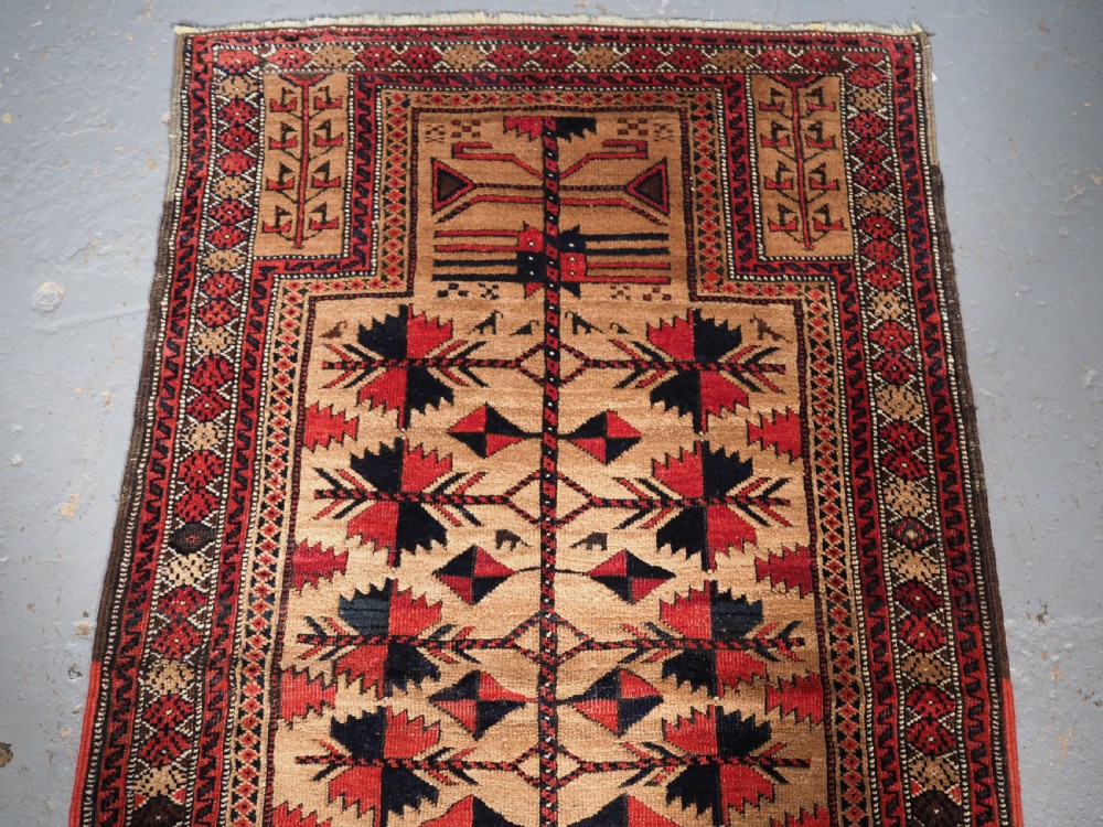 An antique Baluch wool rug featuring a central Tree of Life motif on a camel-coloured ground, framed by geometric red and black patterned borders. View III