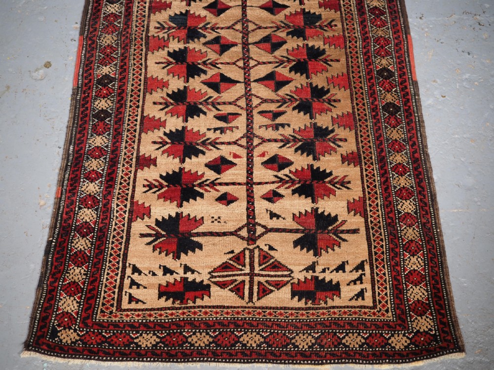 An antique Baluch wool rug featuring a central Tree of Life motif on a camel-coloured ground, framed by geometric red and black patterned borders. View IV
