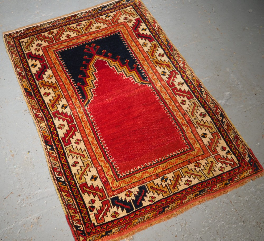 An antique hand-knotted Turkish Konya prayer rug featuring a deep red central stepped mihrab arch bordered by geometric motifs in navy blue, ochre, and cream. View III