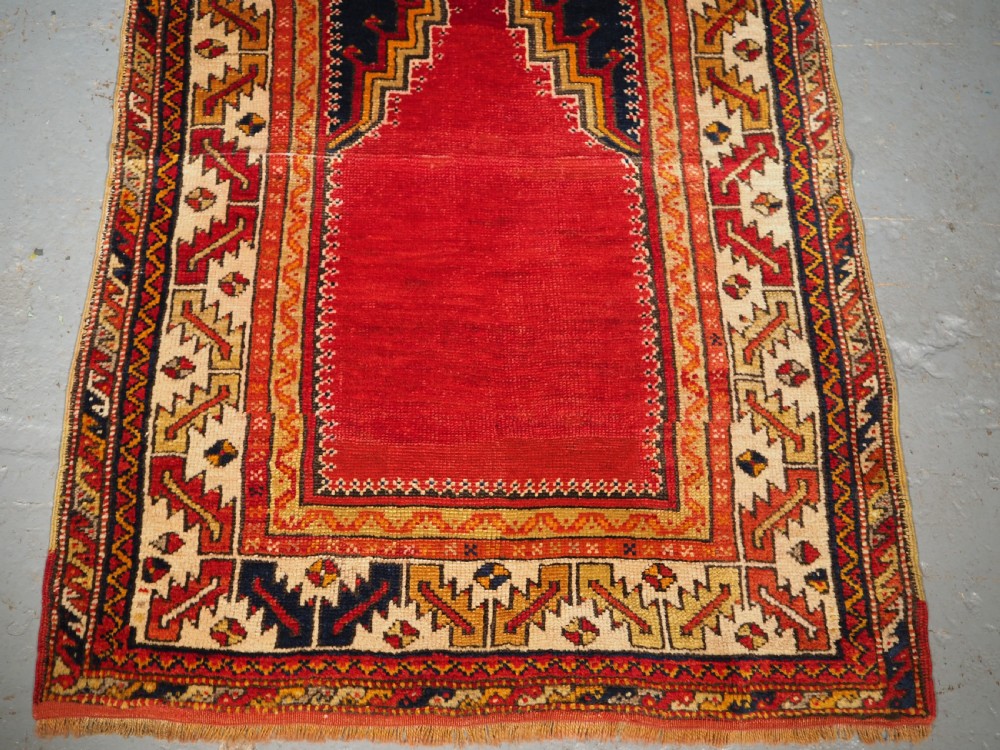 An antique hand-knotted Turkish Konya prayer rug featuring a deep red central stepped mihrab arch bordered by geometric motifs in navy blue, ochre, and cream. View IV