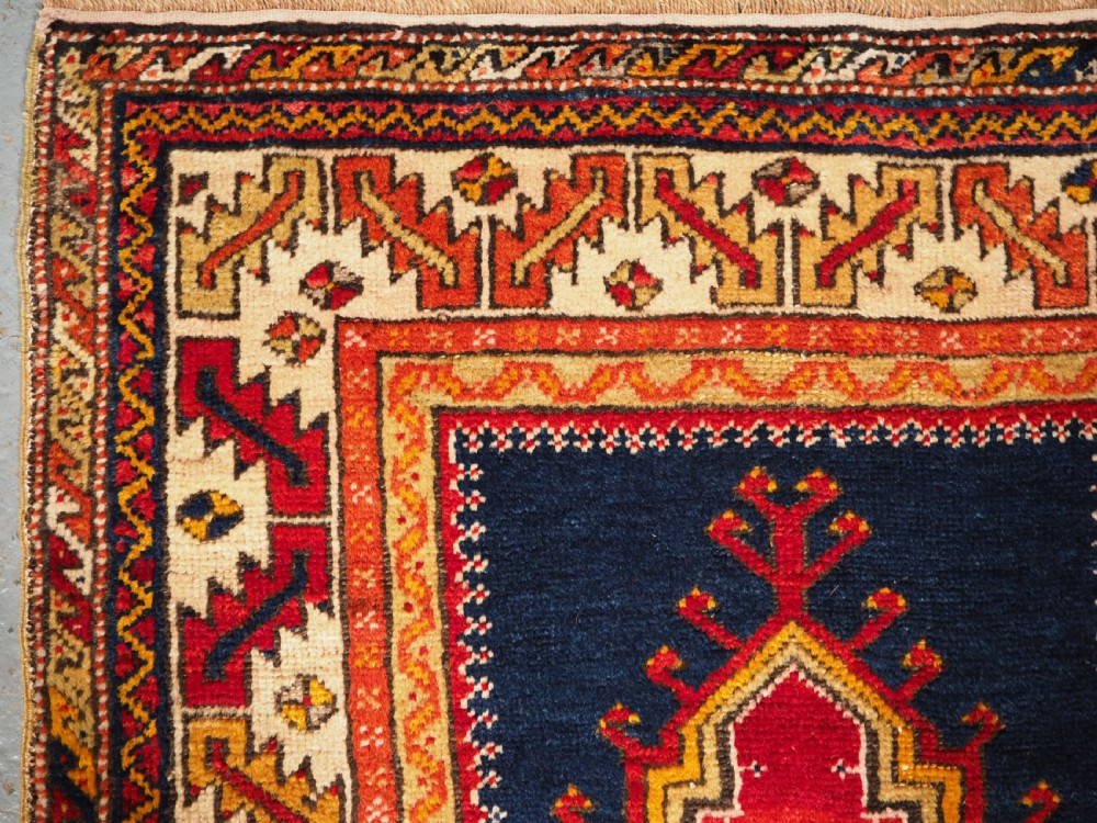 An antique hand-knotted Turkish Konya prayer rug featuring a deep red central stepped mihrab arch bordered by geometric motifs in navy blue, ochre, and cream. View V