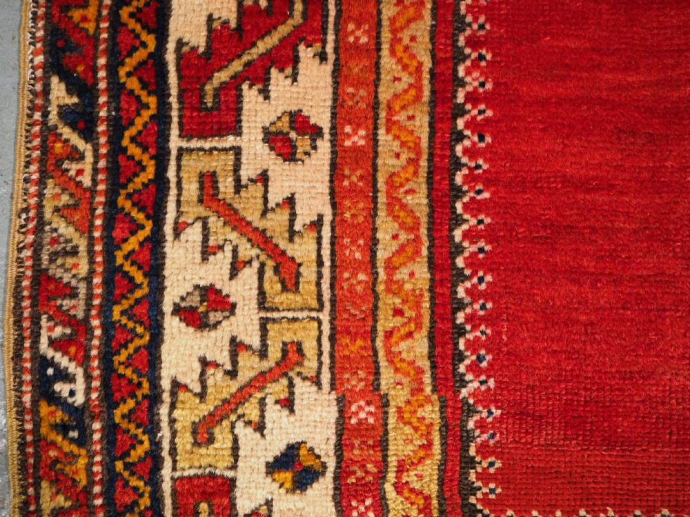 An antique hand-knotted Turkish Konya prayer rug featuring a deep red central stepped mihrab arch bordered by geometric motifs in navy blue, ochre, and cream. View VI