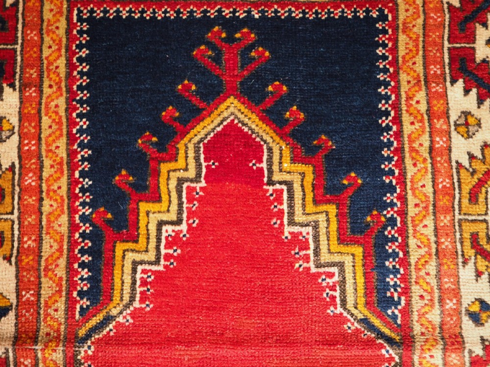 An antique hand-knotted Turkish Konya prayer rug featuring a deep red central stepped mihrab arch bordered by geometric motifs in navy blue, ochre, and cream. View VII