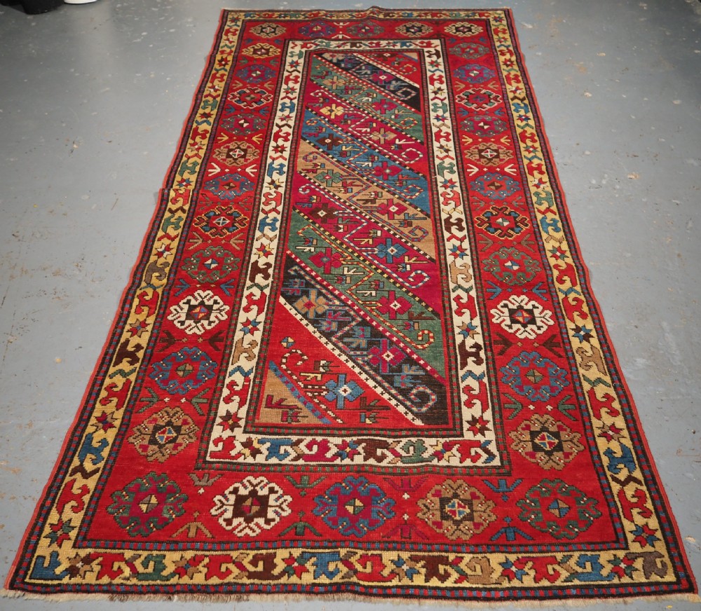 An antique Caucasian Gendje Kazak hand-knotted wool rug featuring diagonal polychrome stripes within a wide geometric border on a red field.