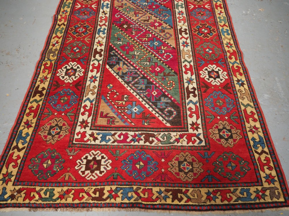 An antique Caucasian Gendje Kazak hand-knotted wool rug featuring diagonal polychrome stripes within a wide geometric border on a red field. View IV