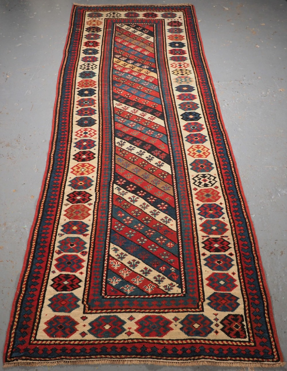 An antique Caucasian Gendje wool runner rug featuring a central field with diagonal stripes in red, blue, and cream surrounded by geometric borders.