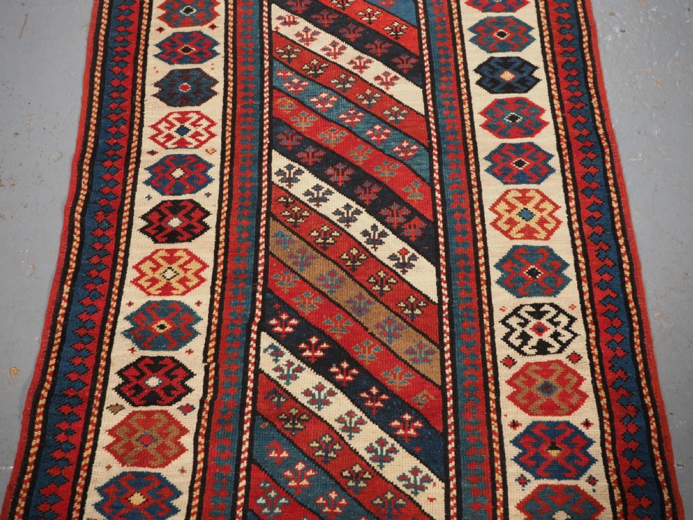 An antique Caucasian Gendje wool runner rug featuring a central field with diagonal stripes in red, blue, and cream surrounded by geometric borders. View IV