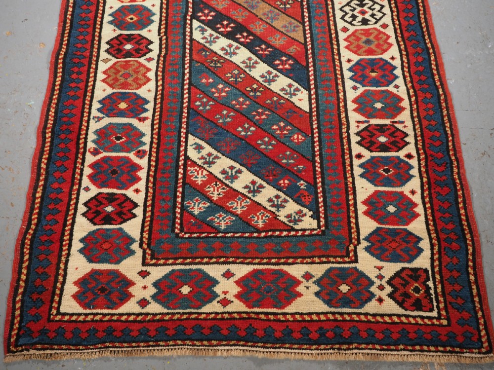 An antique Caucasian Gendje wool runner rug featuring a central field with diagonal stripes in red, blue, and cream surrounded by geometric borders. View V