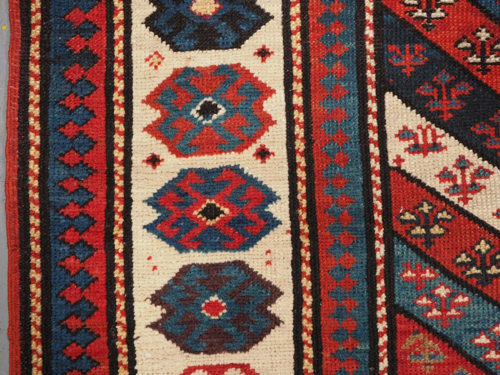 An antique Caucasian Gendje wool runner rug featuring a central field with diagonal stripes in red, blue, and cream surrounded by geometric borders. View VII