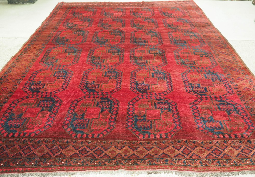 A large hand-knotted wool Afghan carpet featuring a deep red field patterned with repeating dark blue octagonal gul motifs and a geometric border.
