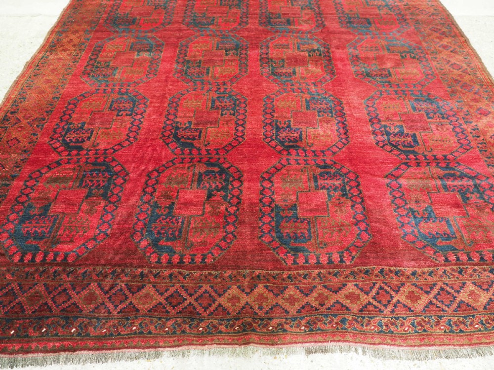 A large hand-knotted wool Afghan carpet featuring a deep red field patterned with repeating dark blue octagonal gul motifs and a geometric border. View VI
