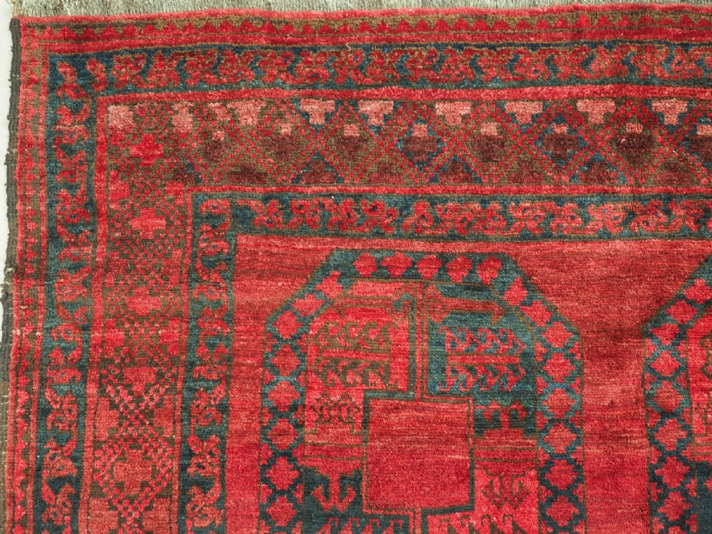 A large hand-knotted wool Afghan carpet featuring a deep red field patterned with repeating dark blue octagonal gul motifs and a geometric border. View VII