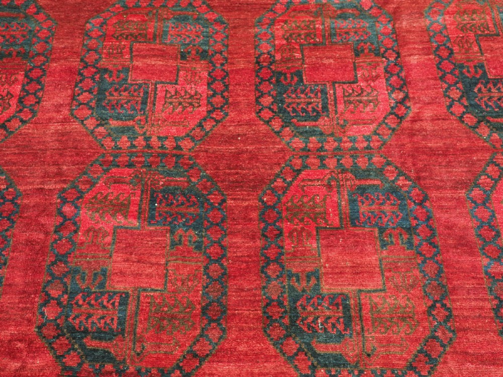 A large hand-knotted wool Afghan carpet featuring a deep red field patterned with repeating dark blue octagonal gul motifs and a geometric border. View IX