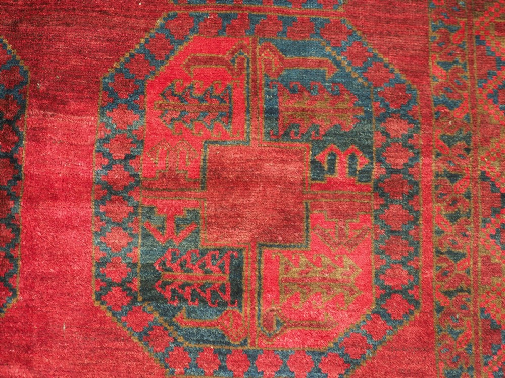 A large hand-knotted wool Afghan carpet featuring a deep red field patterned with repeating dark blue octagonal gul motifs and a geometric border. View X