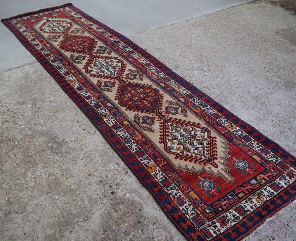 An antique Persian Sarab runner rug featuring repeating geometric diamond medallions on a camel beige central field with terracotta red and navy borders. View II