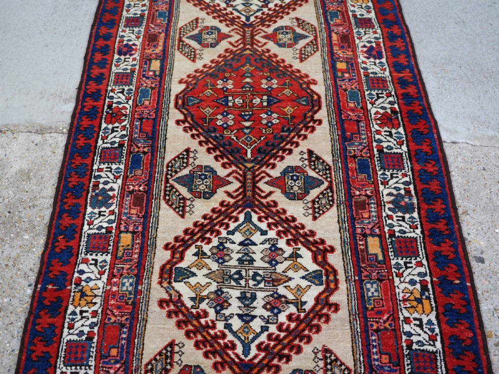 An antique Persian Sarab runner rug featuring repeating geometric diamond medallions on a camel beige central field with terracotta red and navy borders. View IV