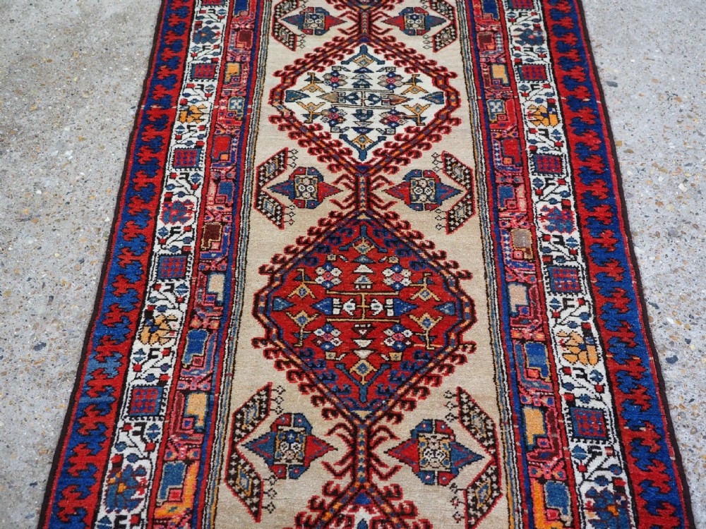 An antique Persian Sarab runner rug featuring repeating geometric diamond medallions on a camel beige central field with terracotta red and navy borders. View V