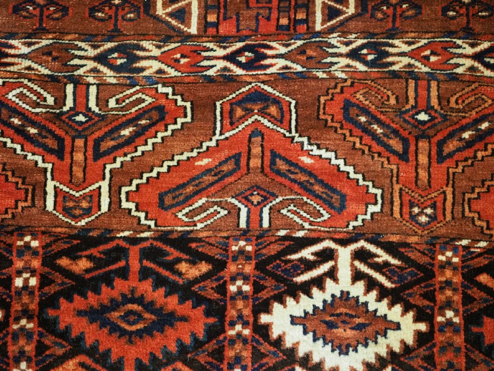 An antique Yomut Turkmen Ensi wool rug featuring a four-part central field design, geometric borders, and an elem end panel in deep red, brown, and ivory. View X