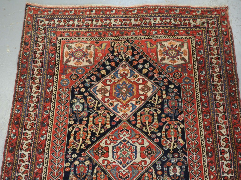 A hand-knotted wool rug featuring three diamond medallions on a dark navy field surrounded by geometric floral borders in red, ivory, and blue. View III
