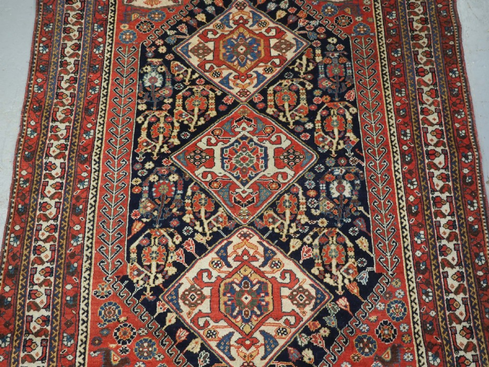 A hand-knotted wool rug featuring three diamond medallions on a dark navy field surrounded by geometric floral borders in red, ivory, and blue. View IV