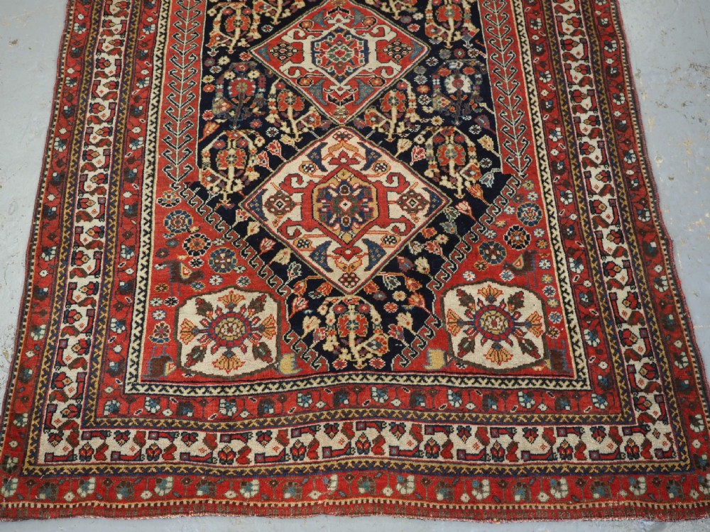 A hand-knotted wool rug featuring three diamond medallions on a dark navy field surrounded by geometric floral borders in red, ivory, and blue. View V