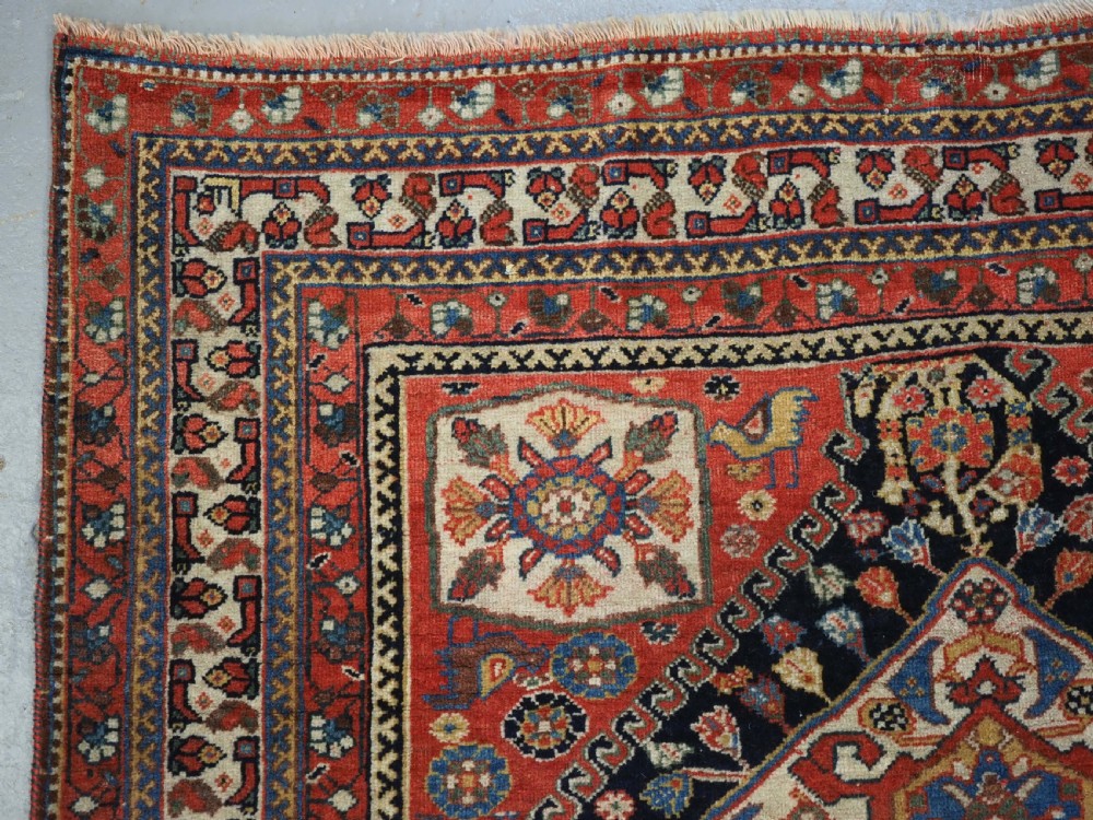 A hand-knotted wool rug featuring three diamond medallions on a dark navy field surrounded by geometric floral borders in red, ivory, and blue. View VI