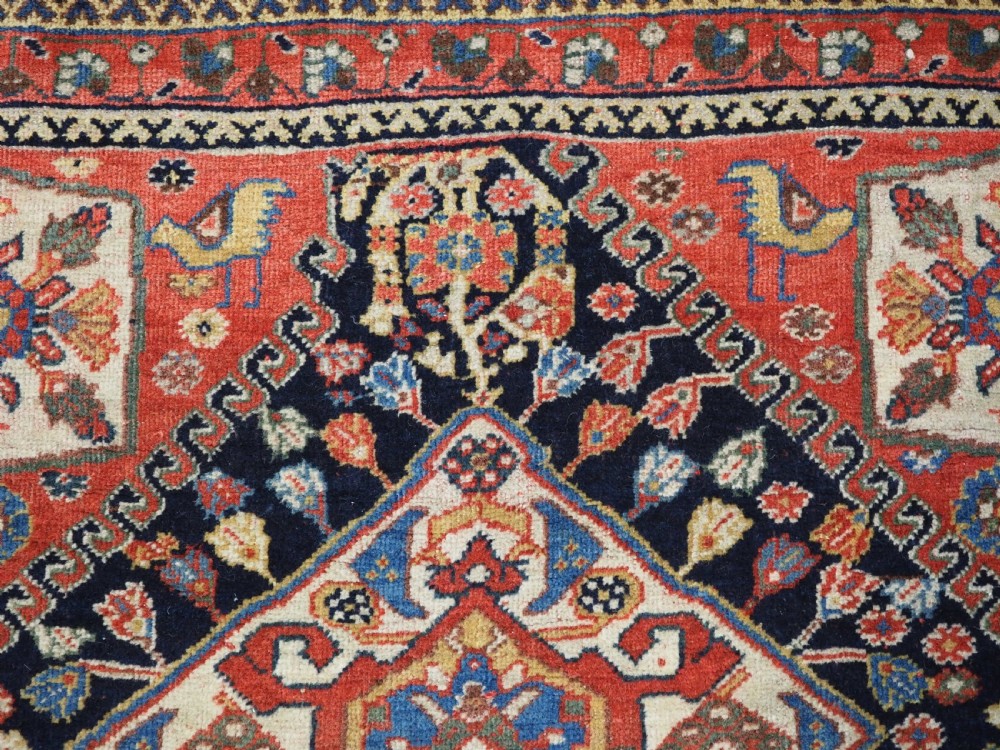 A hand-knotted wool rug featuring three diamond medallions on a dark navy field surrounded by geometric floral borders in red, ivory, and blue. View IX